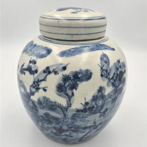 May include: A round ceramic jar with a matching lid, featuring a white base decorated with blue hand-painted illustrations of pine trees, branches, and landscape elements. The lid has horizontal blue line accents around its circumference.
