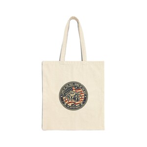 May include: A beige canvas tote bag featuring a circular graphic in the center. The design includes a cartoon spider over a United States flag, surrounded by the text "LAND OF THE FREE, HOME OF THE BUG" and "THE CRITTER DIARIES" in a rope border.