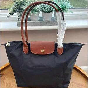 May include: A black nylon tote bag featuring brown leather handles and a brown leather flap closure with a gold-tone snap button. One handle is wrapped in protective white paper. The bag has a zip closure with a small gold-tone circular pull.