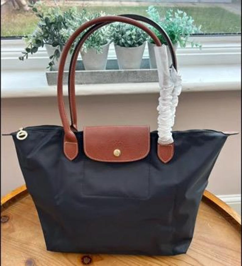 May include: A black nylon tote bag featuring brown leather handles and a brown leather flap closure with a gold-tone snap button. One handle is wrapped in protective white paper. The bag has a zip closure with a small gold-tone circular pull.