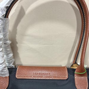 May include: A black nylon handbag featuring brown leather trim and handles. The leather flap is embossed with the text LONGCHAMP, VINTAGE SHOPPING, and MODELE DEPOSE. One handle is partially wrapped in protective white tissue paper for storage.
