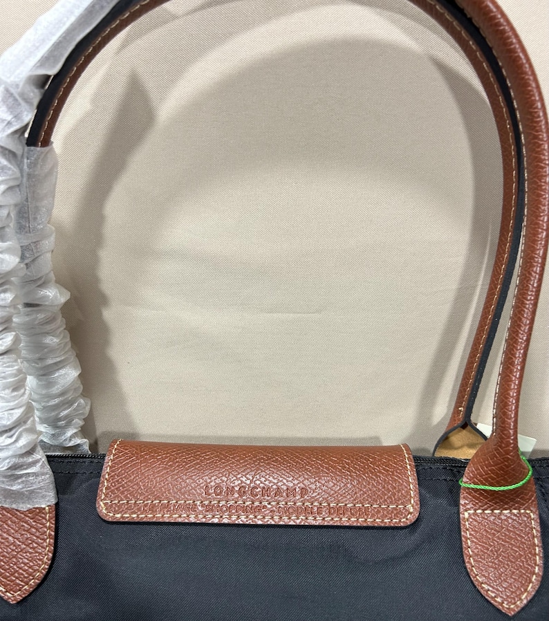 May include: A black nylon handbag featuring brown leather trim and handles. The leather flap is embossed with the text LONGCHAMP, VINTAGE SHOPPING, and MODELE DEPOSE. One handle is partially wrapped in protective white tissue paper for storage.