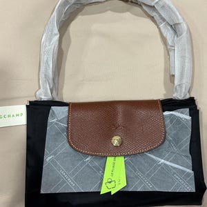 May include: A black Longchamp handbag with a brown leather flap featuring a gold-tone snap button. The handle is wrapped in protective white paper. A lime green tag labelled LE PLIAGE GREEN is attached, and the bag includes branded tissue paper.