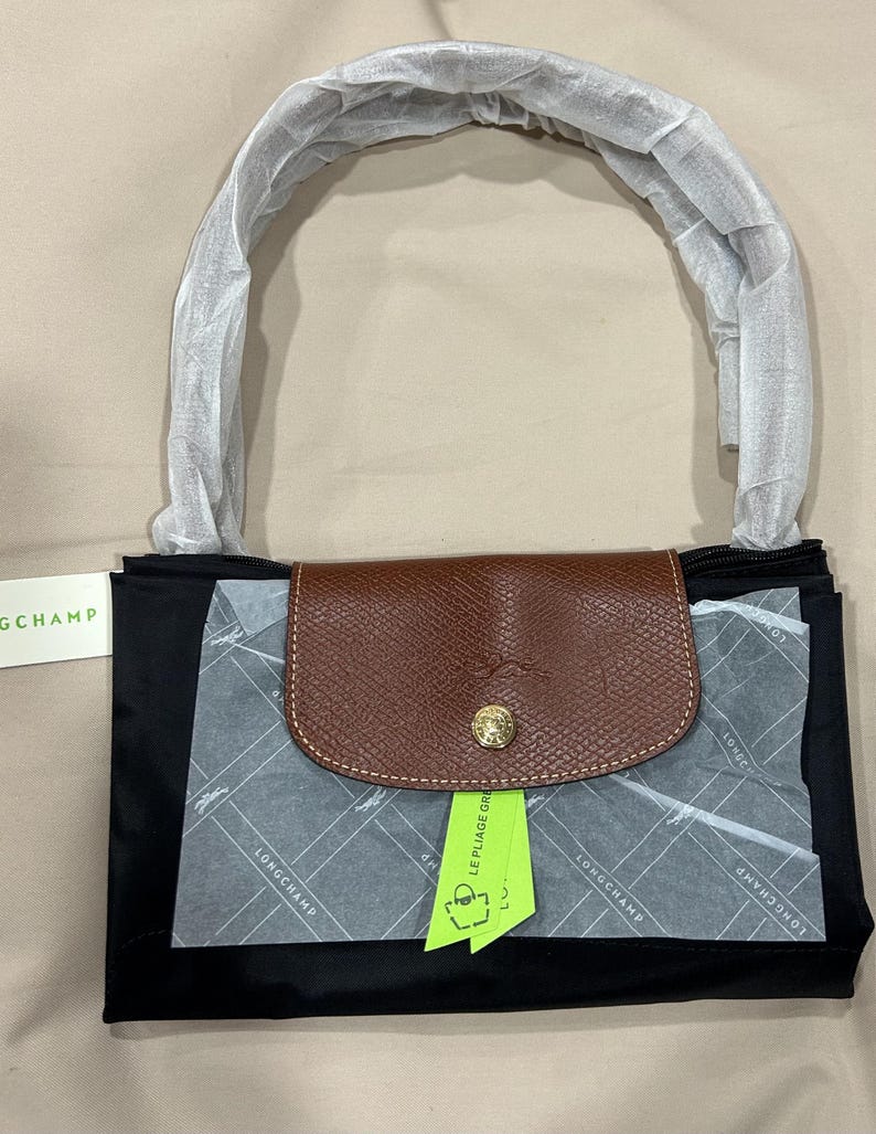 May include: A black Longchamp handbag with a brown leather flap featuring a gold-tone snap button. The handle is wrapped in protective white paper. A lime green tag labelled LE PLIAGE GREEN is attached, and the bag includes branded tissue paper.