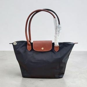 May include: A black nylon tote bag featuring brown leather handles and a brown leather flap closure with a gold-tone snap button. One handle is partially wrapped in protective white plastic. The bag has a zip closure with a gold-tone pull tab.