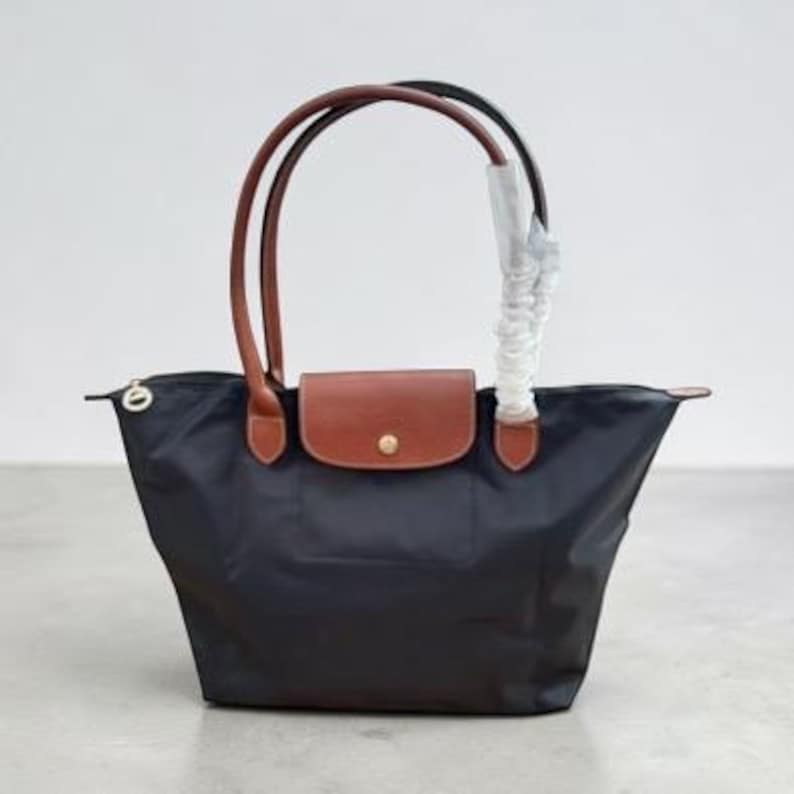 May include: A black nylon tote bag featuring brown leather handles and a brown leather flap closure with a gold-tone snap button. One handle is partially wrapped in protective white plastic. The bag has a zip closure with a gold-tone pull tab.