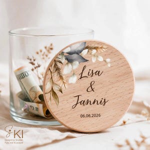 May include: A clear glass jar with a round wooden lid featuring a printed botanical design of leaves and pampas grass. The lid is engraved with the names Lisa & Jannis and the date 06.06.2026. Inside the jar, a rolled-up US dollar bill is visible.