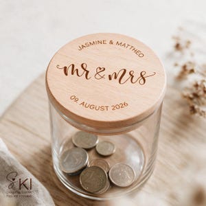 May include: A clear glass jar with a round wooden lid engraved with the text JASMINE & MATTHEW, mr & mrs, and 09. AUGUST 2026. The jar contains several metallic coins visible through the transparent glass walls of the container.