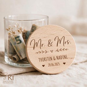May include: A clear glass jar containing rolled-up paper currency, topped with a round wooden lid engraved with the text Mr. & Mrs., the names THORSTEN & NADINE, the date 20.06.2025, and decorative leaf and heart motifs.