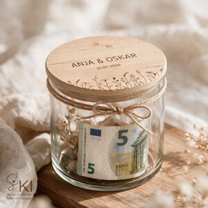 May include: A clear glass jar with a round wooden lid featuring the engraved text ANJA & OSKAR and 31.05.2026, decorated with a floral line art pattern. A jute twine bow is tied around the neck, and a five euro banknote is visible inside.