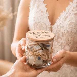 May include: A clear glass jar with a round wooden lid featuring the engraved text Mr. & Mrs. THORSTEN & NADINE 20.06.2025. The jar is wrapped with a natural jute twine bow and contains several folded Euro banknotes inside the transparent glass container.