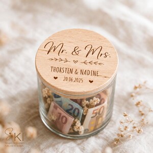 May include: A clear glass jar with a round wooden lid engraved with the text Mr. & Mrs., the names THORSTEN & NADINE, and the date 20.06.2025. The lid features decorative leaf and heart motifs. Inside the jar are folded banknotes and dried flowers.
