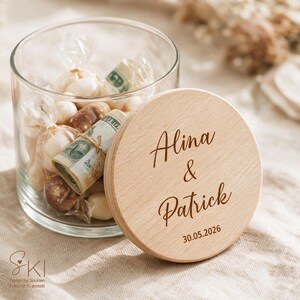 May include: A clear glass jar filled with individually wrapped candies and rolled-up US dollar bills. The jar has a round wooden lid engraved with the names Alina & Patrick and the date 30.05.2026 in a dark brown script and sans-serif font.