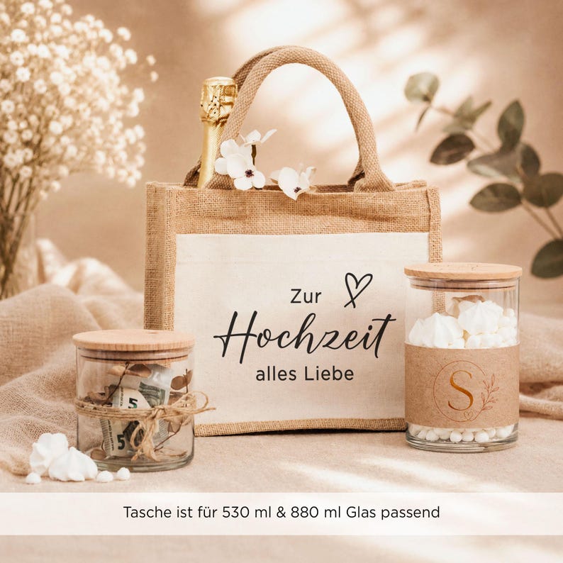 May include: A jute gift bag with the text Zur Hochzeit alles Liebe and a heart icon, containing a champagne bottle. Two glass jars with wooden lids are placed in front, one filled with paper money and the other with white meringues.