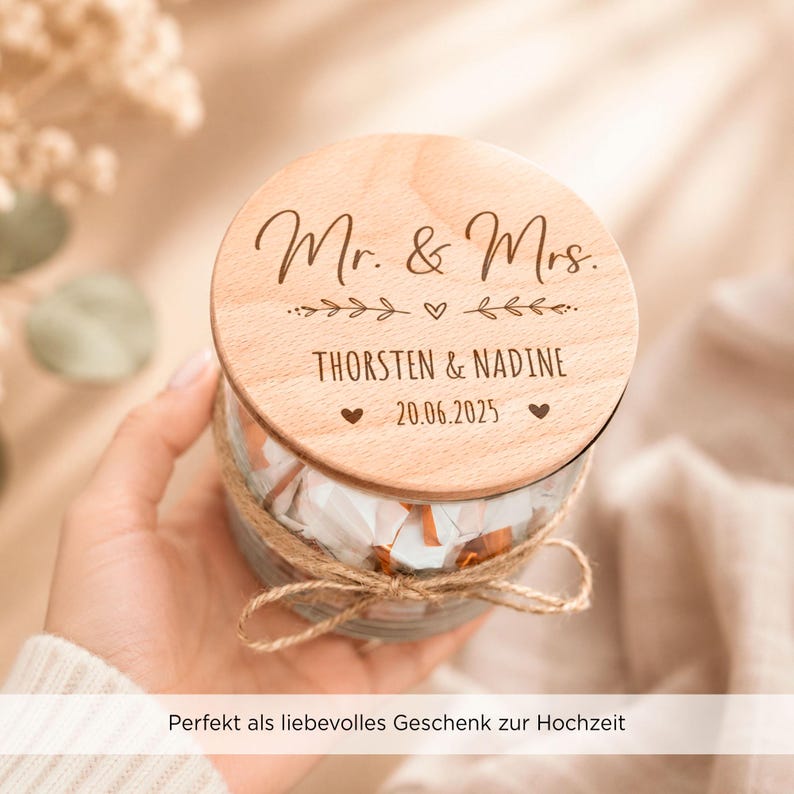 May include: A clear glass jar with a round wooden lid featuring the engraved text Mr. & Mrs., the names THORSTEN & NADINE, and the date 20.06.2025. The lid is decorated with leaf motifs and small hearts. A jute twine bow is tied around the jar neck.