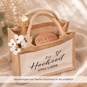 May include: A small jute tote bag with two handles and a white front pocket featuring the black text Zur Hochzeit alles Liebe and a heart icon. Inside the bag is a champagne bottle, a round wooden lid with Mr. & Mrs. text, and white flowers.