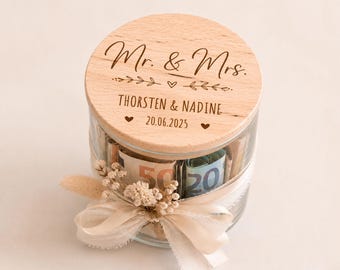 Könnte beinhalten: Ein klares Glas mit gerollten Banknoten, bedeckt mit einem Holzdeckel. Der Deckel ist mit "Mr. & Mrs.", Namen und einem Datum graviert. Ein Band und Trockenblumen schmücken das Glas, was auf eine Hochzeit oder einen besonderen Anlass hindeutet.