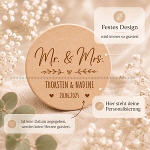 May include: A round, light-colored wooden disc featuring laser-engraved text. The top section reads Mr. & Mrs. with a small heart and leaf design. The bottom section includes the names THORSTEN & NADINE and the date 20.06.2025 with two hearts.