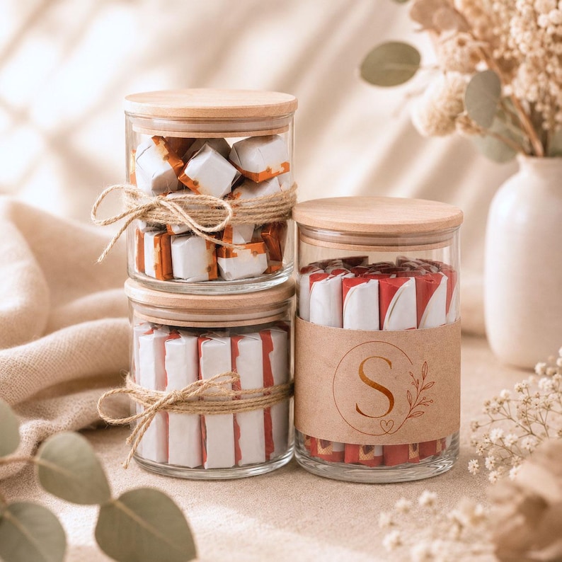 May include: Two clear glass jars with light wood lids filled with individually wrapped candies. The top jar and the bottom left jar are tied with natural jute twine. The bottom right jar features a tan paper label with a large letter S.