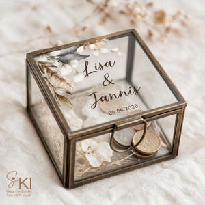 Könnte beinhalten: Eine quadratische Glasbox mit einem messingfarbenen Metallrahmen. Der Deckel zeigt ein botanisches Motiv mit den Namen Lisa & Jannis und dem Datum 06.06.2026. Im Inneren der Box liegen mehrere Metallmünzen auf einem Bett aus getrockneten Blumen.