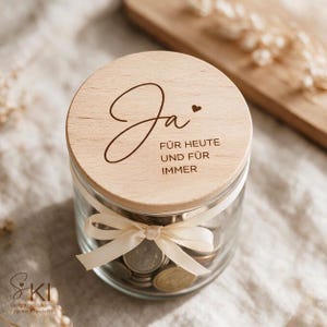 May include: A clear glass jar filled with metal coins, featuring a round light-colored wooden lid engraved with the German text Ja, FÜR HEUTE UND FÜR IMMER and a small heart symbol. A cream-colored ribbon is tied around the neck of the glass jar.