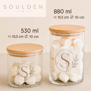May include: Two clear glass storage jars with light wood lids. The smaller jar holds 530 ml, H: 10.5 cm, &Oslash;: 10 cm. The larger jar holds 880 ml, H: 15.5 cm, &Oslash;: 10 cm. Each jar features a circular monogram with the letter S and a floral sprig.