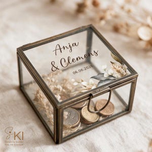 May include: A square glass box with a dark metal frame and a hinged lid. The lid features the printed text Anja & Clemens and the date 06.06.2026. Inside the box are several metallic coins and dried floral elements in neutral tones.