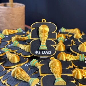 May include: A collection of black and gold 3D-printed ornaments shaped like the number 26, featuring a gold trophy icon with green accents and the white text #1 DAD. Each ornament has a small loop at the top for hanging, with a textured surface.