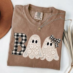 May include: A brown short-sleeved t-shirt featuring a graphic of the letter B in black and white buffalo check, followed by two white lace ghosts. One ghost wears a black and white buffalo check bow. The shirt has a Comfort Colors brand label.