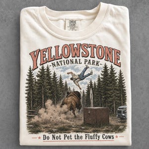Könnte beinhalten: Ein cremefarbenes T-Shirt mit einer Grafik, die eine Person zeigt, die von einem Bison in einem Wald hochgeschleudert wird. Der Text lautet YELLOWSTONE NATIONAL PARK und Do Not Pet the Fluffy Cows. Das Design zeigt Kiefern, eine Holzkiste und ein Fahrzeug.