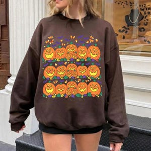 Könnte beinhalten: Ein dunkelbraunes Langarm-Sweatshirt mit einem Grafikdruck von drei Reihen orangefarbener Kürbisse, kleinen gelben Schmetterlingen, violetten Fledermäusen und Zuckermais, die auf der Vorderseite des Kleidungsstücks verteilt sind.