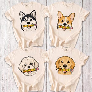 May include: Four cream-colored t-shirts, each featuring a graphic illustration of a different dog breed—a husky, a corgi, a doodle, and a golden retriever—holding a yellow pencil with colorful star and apple patterns in its mouth.