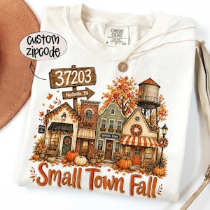 Pode incluir: Uma t-shirt cor de creme com um gráfico de uma rua de cidade pequena no outono, com abóboras, folhagem de outono, uma torre de água e uma placa com 37203 Nashville, TN. O texto Small Town Fall está impresso em laranja na parte inferior.