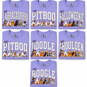 May include: Seven light purple t-shirts featuring various dog breeds in Halloween costumes. The shirts display text including LABRACADABRA, PITBOO, HALLOWEENIE, DOODLE, GHOULDEN, and BOOGLE, with illustrations of dogs dressed as ghosts, pumpkins, and skeletons.