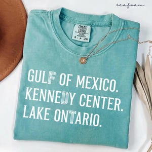 May include: A seafoam green short-sleeve t-shirt with a crew neckline. The shirt features white text that reads GULF OF MEXICO. KENNEDY CENTER. LAKE ONTARIO. A thin gold-tone chain necklace with a small circular pendant rests on the fabric.