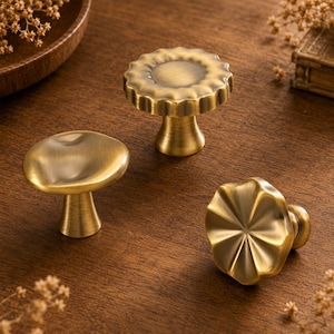 May include: Three brass-toned metal cabinet knobs with a brushed finish. One knob features a smooth, rounded mushroom-like top, another has a scalloped edge, and the third displays a radial, sunburst-like pattern on its circular face.