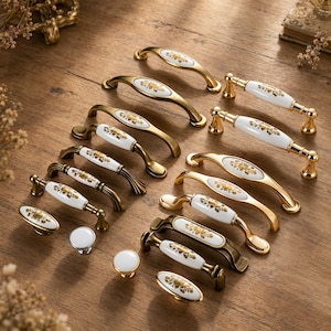 Può includere: Una collezione di ferramenta decorativa per mobili, tra cui maniglie e pomelli. La ferramenta presenta un centro in ceramica bianca con un motivo floreale dorato e dettagli in metallo dorato o bronzo. Vengono mostrate varie forme e dimensioni.