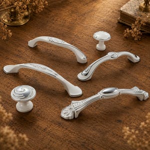 May include: A collection of cabinet hardware including three arched drawer pulls and two round knobs. The hardware features a white finish with polished silver metallic accents, showcasing various embossed patterns and smooth, curved surfaces.