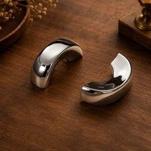 May include: Two polished silver-toned metal objects with a smooth, curved, crescent-like shape. Each piece features a reflective, high-gloss finish that captures light, creating bright highlights across the rounded, solid, and arching surfaces.