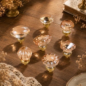 May include: A collection of seven cabinet knobs featuring clear, light amber-tinted glass tops in various geometric and faceted shapes, each mounted on a circular brushed brass base. The set includes round, square, and textured glass designs.