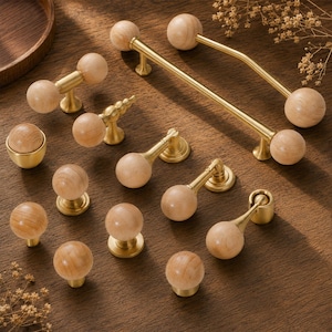 May include: A collection of cabinet hardware featuring light wood spheres paired with brushed brass metal bases. The set includes various drawer pulls, knobs, and drop handles, showcasing a combination of natural wood grain and metallic finishes.