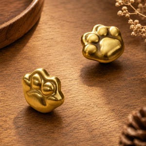 Pode incluir: Dois puxadores de armário em metal dourado com formato de pegadas de animais. Cada puxador apresenta uma almofada central elevada e quatro almofadas menores com um acabamento escovado suave. As ferragens são projetadas para móveis ou portas.