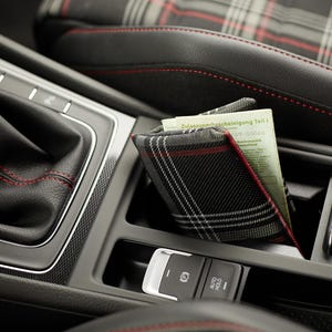 Può includere: Un portadocumenti in tessuto a quadri neri, bianchi e rossi contenente un documento di immatricolazione tedesco con l'etichetta Zulassungsbescheinigung Teil I, posizionato nella console centrale di un'auto in pelle nera con cuciture rosse.