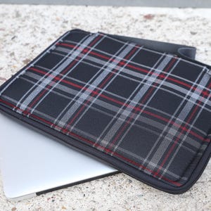 Pode incluir: Uma capa retangular para portátil com um padrão de xadrez preto, cinzento e vermelho. A capa tem um rebordo com fecho de correr preto e está colocada sobre um portátil prateado, exibindo um design estruturado com superfície plana e cantos arredondados.