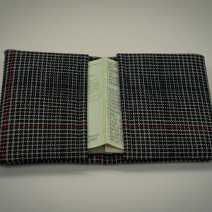 May include: A rectangular wallet featuring a black, white, and grey glen plaid pattern with a thin red horizontal stripe. The wallet is open in the centre, revealing a folded document with visible printed text inside the central compartment.