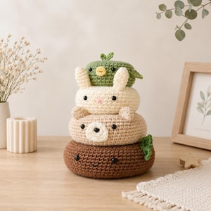 May include: A stack of four crocheted plush toys in the shape of animals. The bottom is a dark brown bear, followed by a tan bear, a cream-coloured rabbit, and a green bird on top. Each piece is round and features black bead eyes and stitched details.