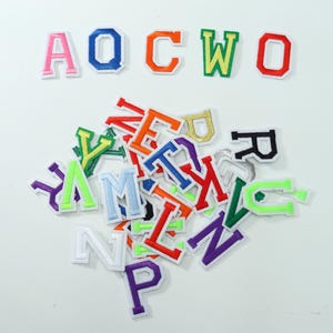 Può includere: Una collezione di toppe con lettere ricamate in stile varsity in vari colori, tra cui rosa, blu, arancione, verde, rosso, viola, giallo e bianco. Le lettere sono disposte con una fila A, O, C, W, O in alto e un mucchio sottostante.