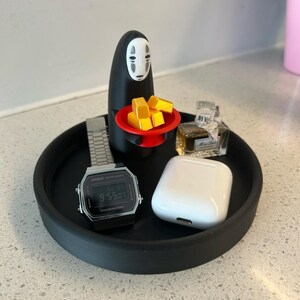 May include: A black tray holds a silver digital watch, a white wireless earbud case, and a small perfume bottle. A black figure with a white face holds a red plate with yellow blocks. The tray sits on a speckled surface.