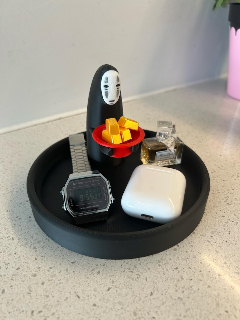 May include: A black tray holds a silver digital watch, a white wireless earbud case, and a small perfume bottle. A black figure with a white face holds a red plate with yellow blocks. The tray sits on a speckled surface.
