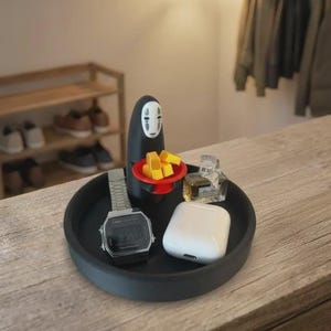 May include: A black round tray holds a digital watch, white wireless earbuds, and small perfume bottles. A black figure with a white face and a red plate with yellow blocks sits on the tray. The tray is on a wooden surface.